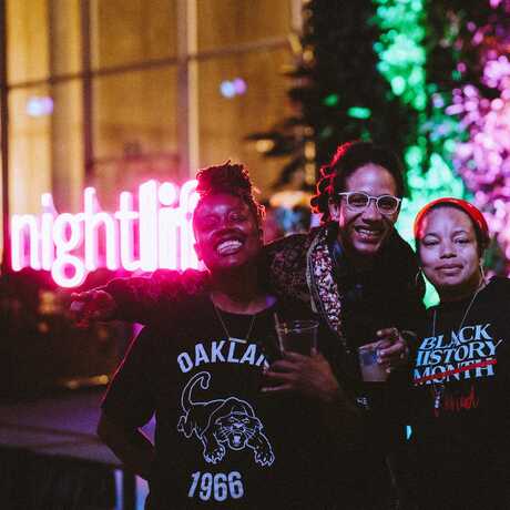 Three friends smile for the camera in a dark room with a neon NightLife sign behind them